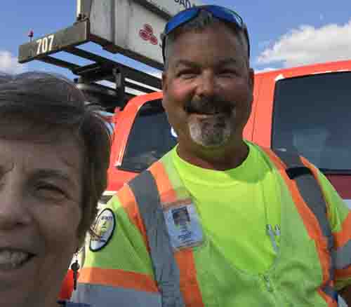 picture shows Dona with a smiling man with a salf-and-pepper beard, wearing a yellow and orange vest, with this truck behind them.